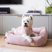 Load image into Gallery viewer, White dog in a pink dog bed on a wooden floor with a TV and books in the background.
